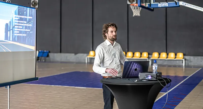 A speaker gives a presentation next to a large screen on the basketball court