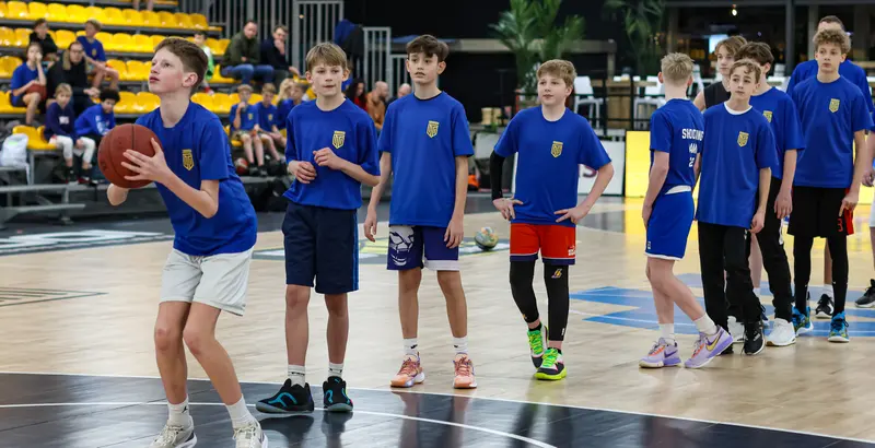 Jonge spelers in blauwe United-shirts staan op een rij in een grote sporthal. De speler voorop mikt op de basket.