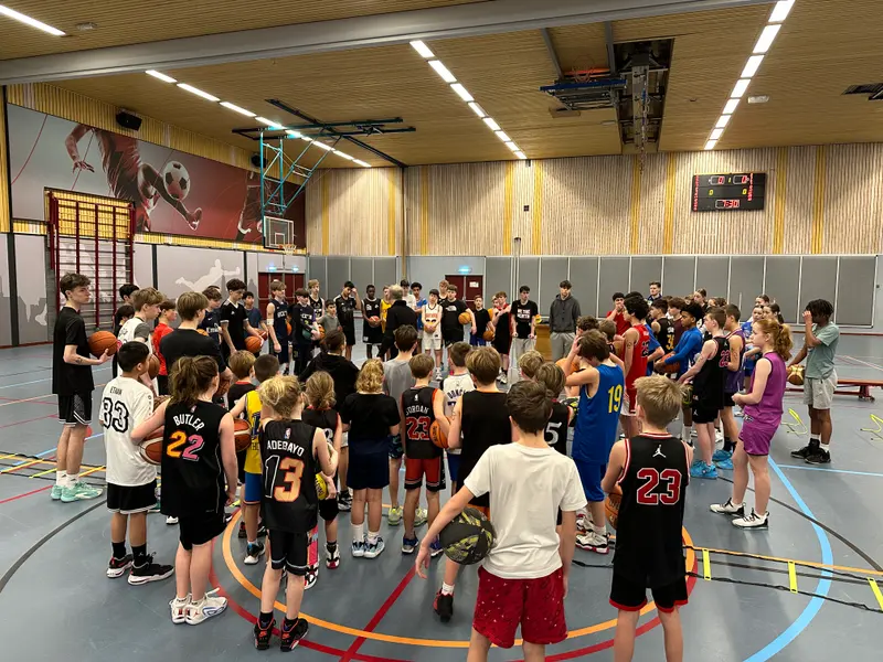 Een grote groep jonge spelers staat in een kring in een gymzaal en luistert naar de uitleg van een trainer, vlak voor een training.