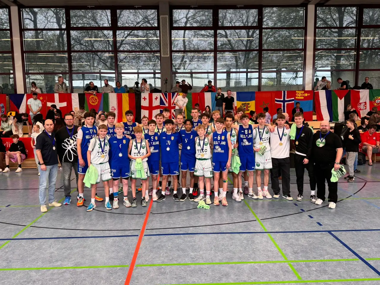 Spelers en begeleiders van United Basketball met hun medailles op het toernooi in Berlijn in 2024, met de vlaggen van de deelnemende landen op de achtergrond.